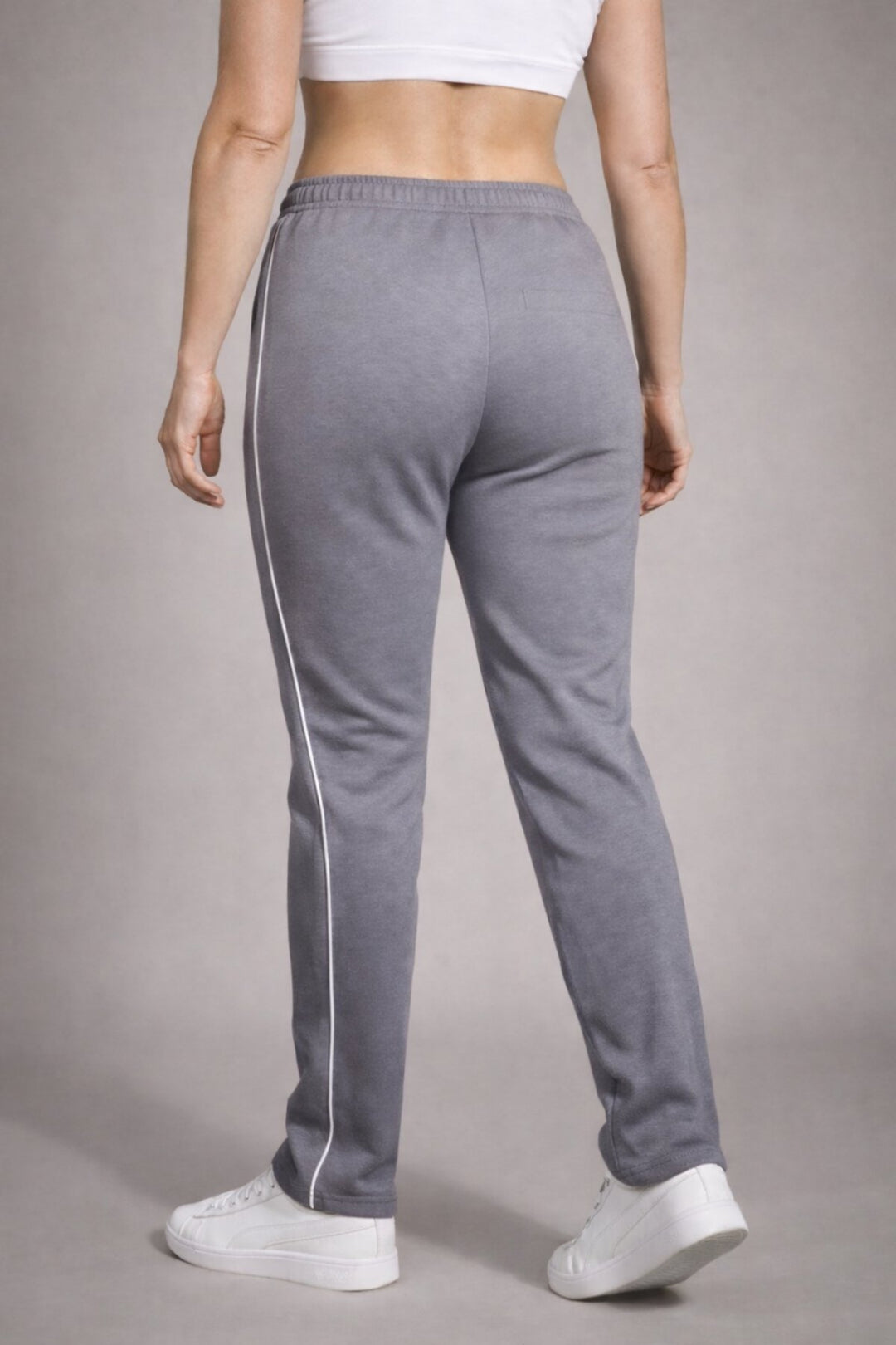 Achteraanzicht van de grijze dames joggingbroek, gedragen door een model, met focus op de pasvorm van de billen, de elastische tailleband en de doorlopende witte zijstreep. Tegen een effen beige achtergrond.