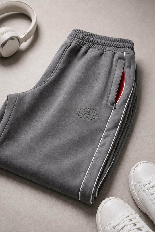 Close-up van de grijze stof van de dames joggingbroek, met focus op het subtiele geborduurde logo op het bovenbeen en de contrasterende witte bies langs de zijkant.