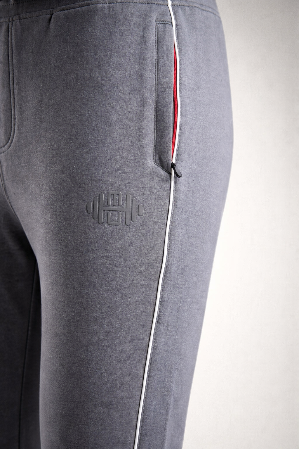 Close-up van de grijze stof van de heren joggingbroek, met focus op het subtiele geborduurde logo op het bovenbeen en de contrasterende witte bies langs de zijkant.