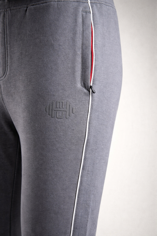Close-up van de grijze stof van de heren joggingbroek, met focus op het subtiele geborduurde logo op het bovenbeen en de contrasterende witte bies langs de zijkant.