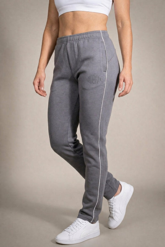 zij-aanzicht van een model dat een grijze joggingbroek voor dames draagt met een witte zijstreep, een subtiel logo op het bovenbeen en zwarte sneakers.