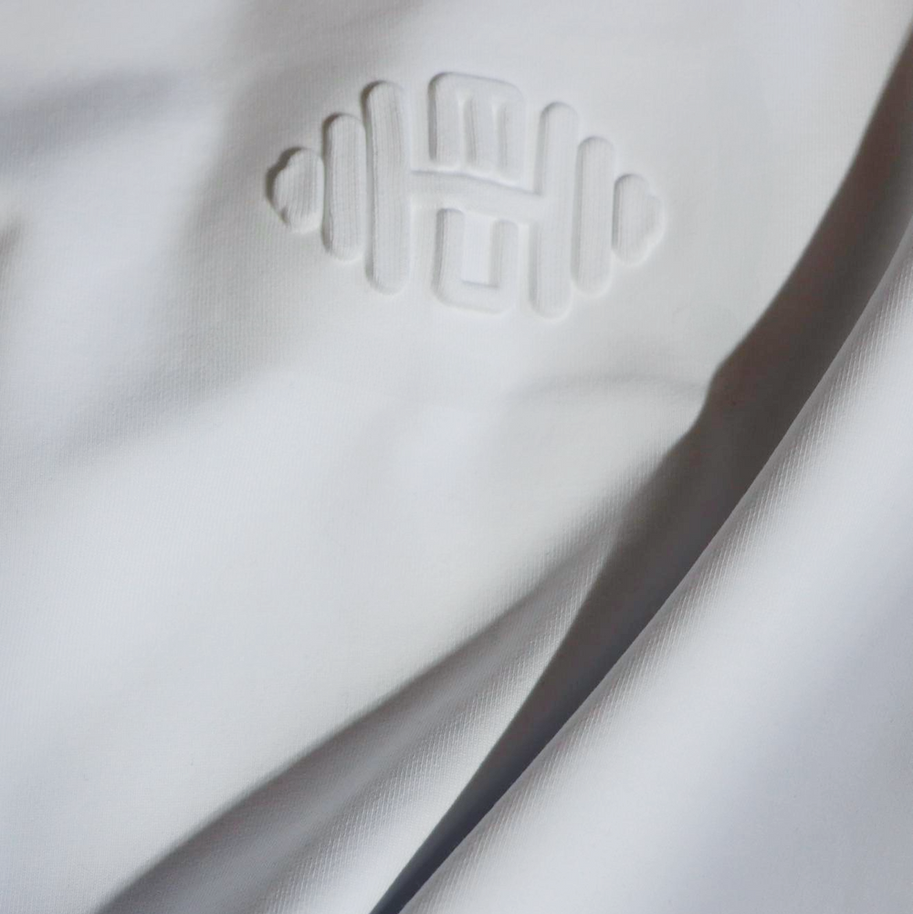 Close-up van een witte oversized T-shirt met een subtiel embossed logo in de vorm van een halter, minimalistisch en luxe afgewerkt.