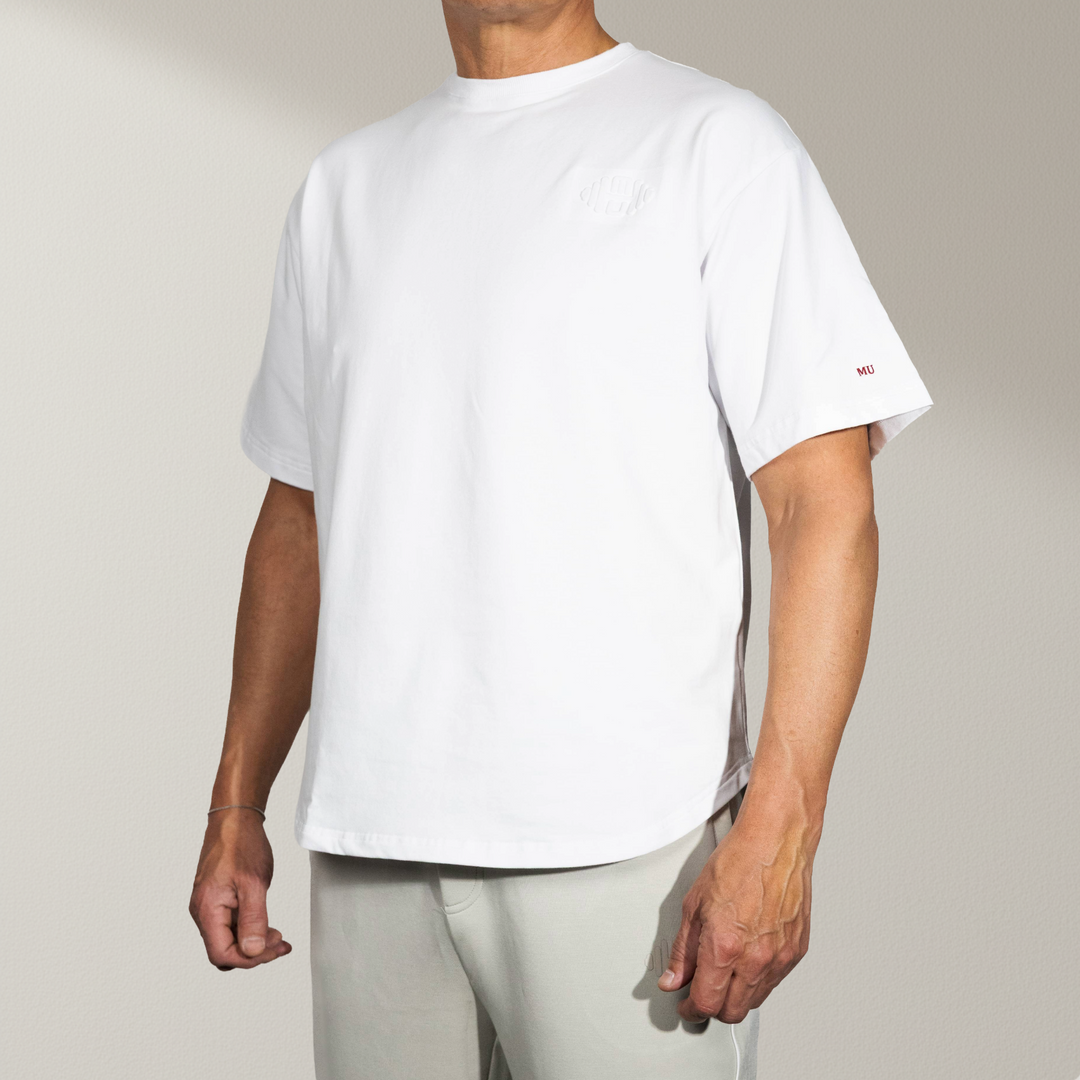 Wit oversized T-shirt met korte mouwen, gedragen door een mannelijk model vanuit de voorkant/schuin. De white tee heeft subtiele rode 'MU' branding op de mouw en een toon-op-toon logo op de borst. Perfect voor een moderne, luxe en comfortabele stijl.