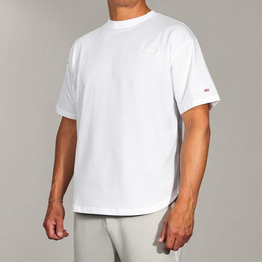 Wit oversized T-shirt met korte mouwen, gedragen door een mannelijk model vanuit de voorkant/schuin. De white tee heeft subtiele rode 'MU' branding op de mouw en een toon-op-toon logo op de borst. Perfect voor een moderne, luxe en comfortabele stijl.