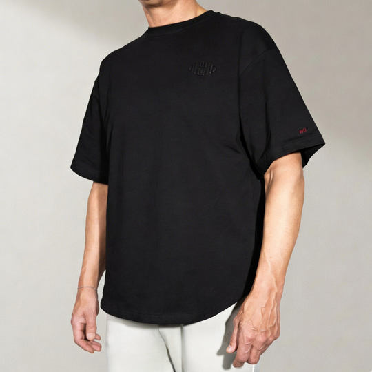 Zwart oversized T-shirt met korte mouwen, gedragen door een mannelijk model vanuit de voorkant/schuin. De black tee heeft subtiele rode 'MU' branding op de mouw en een toon-op-toon logo op de borst. Perfect voor een moderne, luxe en comfortabele stijl.