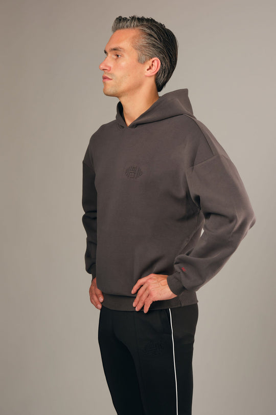 Model draagt Hoodie G2 – luxe grijze oversized en cropped herenhoodie met embossed logo, stijlvolle pasvorm en casual uitstraling